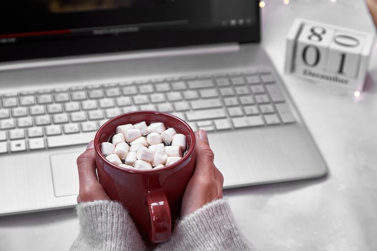 Cozy home office : laptop and red cup with marshmallows close up. Festive working atmosphere with garlands.1st of December. Winter cozy home atmosphere. Top view.